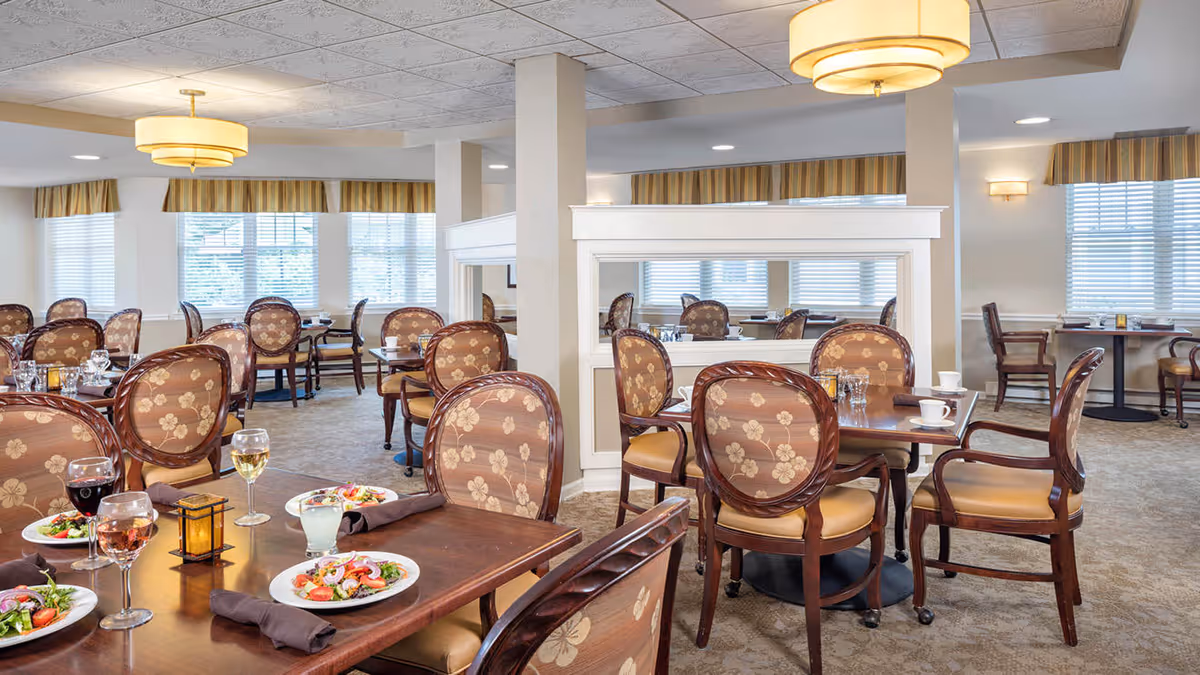 A bright and spacious dining room with multiple wooden tables and upholstered chairs arranged neatly. Each table is set with plates of salad, glasses of wine and water, napkins, and a small decorative lantern. Large windows with striped valances allow natural light to fill the room, and a large ceiling light fixture hangs overhead.
