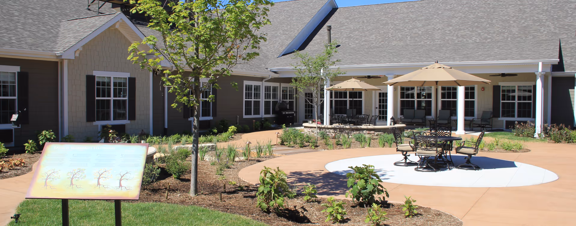 Outdoor courtyard area at Bickford of Tinley Park featuring a circular paved seating area with tables, chairs, and umbrellas. The courtyard is surrounded by a garden with small trees and plants, and the building with multiple windows and a covered patio is visible in the background.