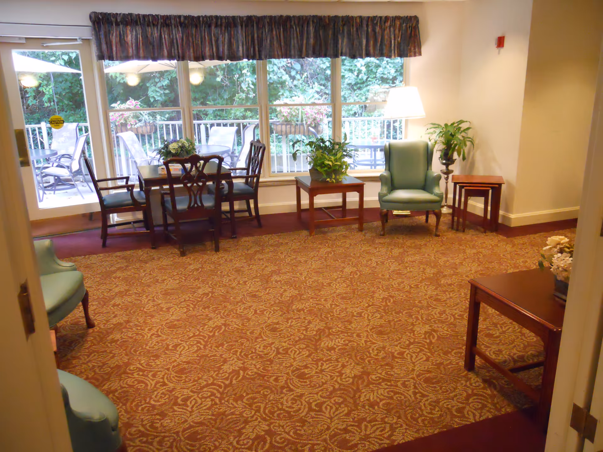 A cozy sitting area in a senior living facility with patterned carpet, green upholstered armchairs, wooden tables with plants and flowers, and a large window showing an outdoor patio with chairs and tables surrounded by greenery.
