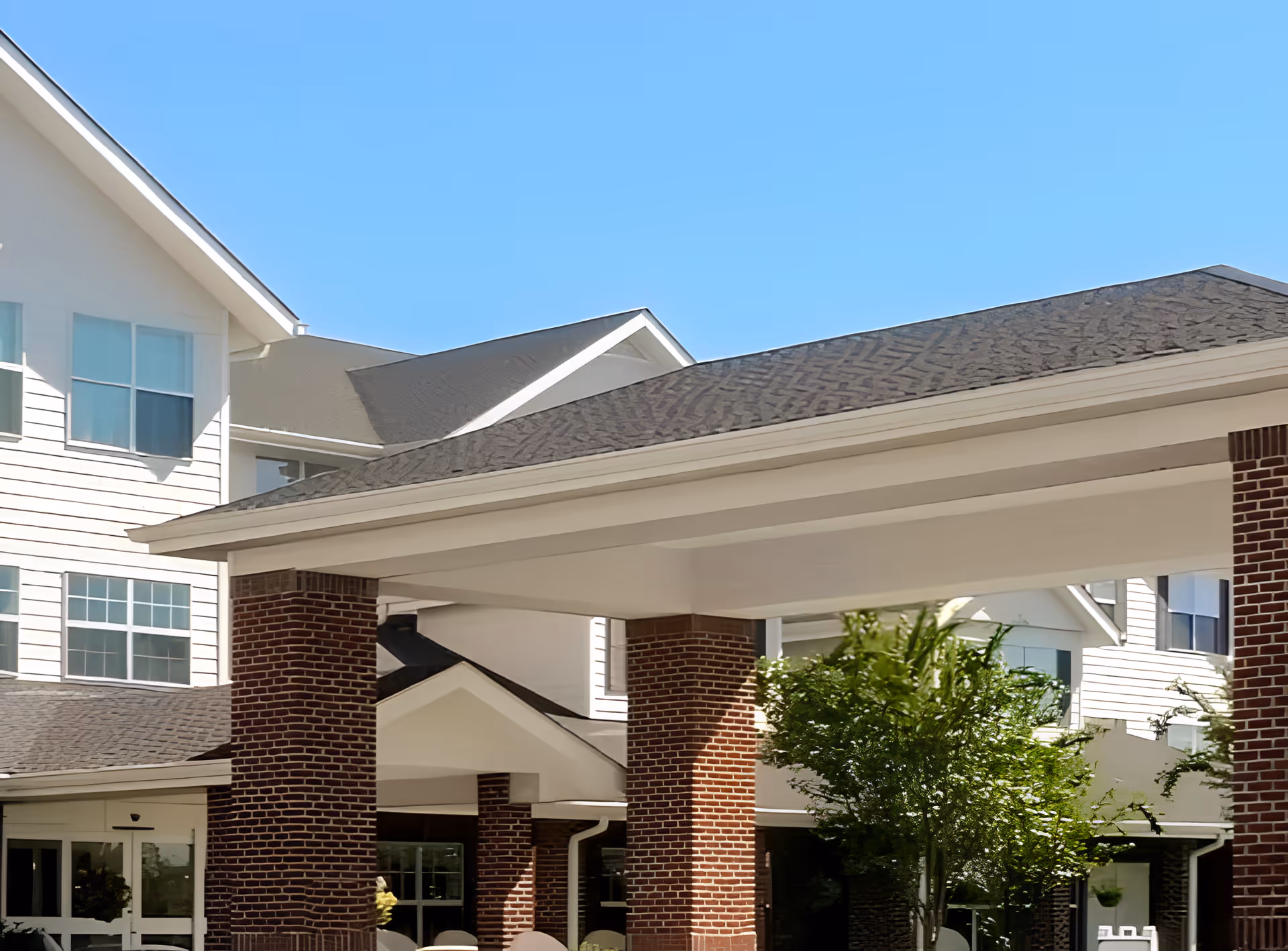 Exterior view of TerraBella Myrtle Beach senior living facility showing a covered entrance with brick pillars, white siding, multiple windows, and a clear blue sky.