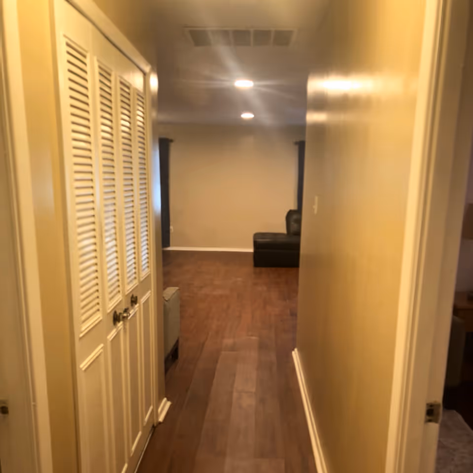View down a hallway with wooden flooring and beige walls. On the left side, there are white louvered closet doors with two knobs. At the end of the hallway, there is a room with a dark-colored sectional sofa and two windows with dark curtains. The ceiling has recessed lighting.