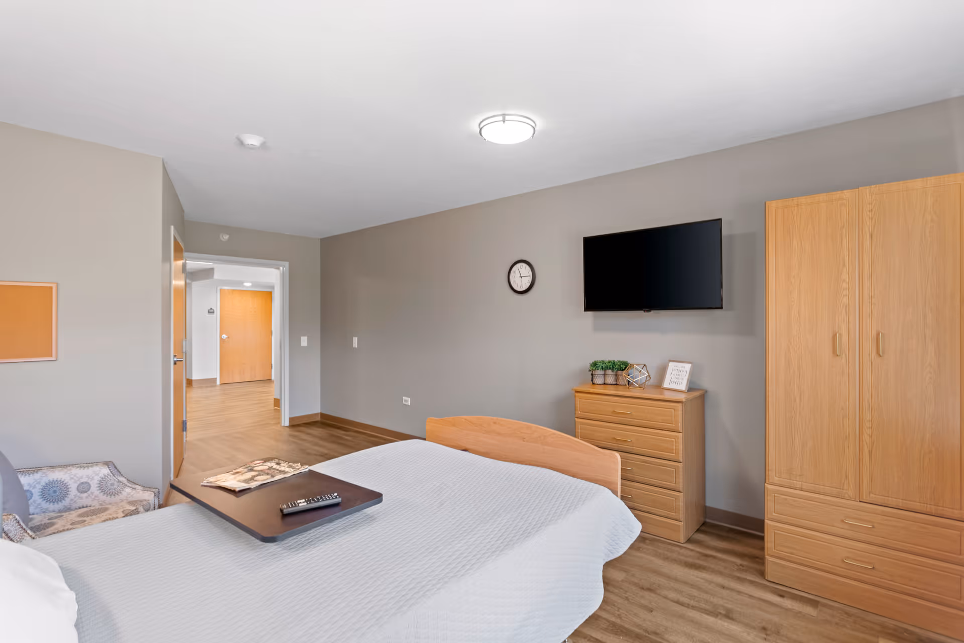 A nursing facility bedroom with a bed covered in a white quilt, a wooden dresser with decorative items, a wall-mounted flat-screen TV, a wooden wardrobe, a patterned armchair, and a small table tray on the bed. The room has light gray walls and wood flooring, with a hallway visible through an open door.