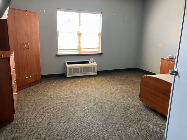 A simple, sparsely furnished room with a window covered by blinds, a wooden wardrobe, a wooden dresser, a wooden bed frame, and a wall-mounted air conditioning unit beneath the window. The walls are painted light gray and the floor is carpeted.
