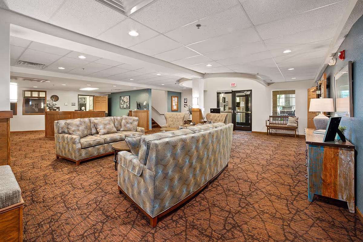 A spacious senior living facility common area with patterned brown carpet, two patterned sofas, two armchairs, a round wooden coffee table, and a wooden reception desk in the background. The walls are painted white and teal, with framed artwork and a lamp on a rustic wooden console table. There is a bench near the glass exit doors and ceiling lights illuminating the room.