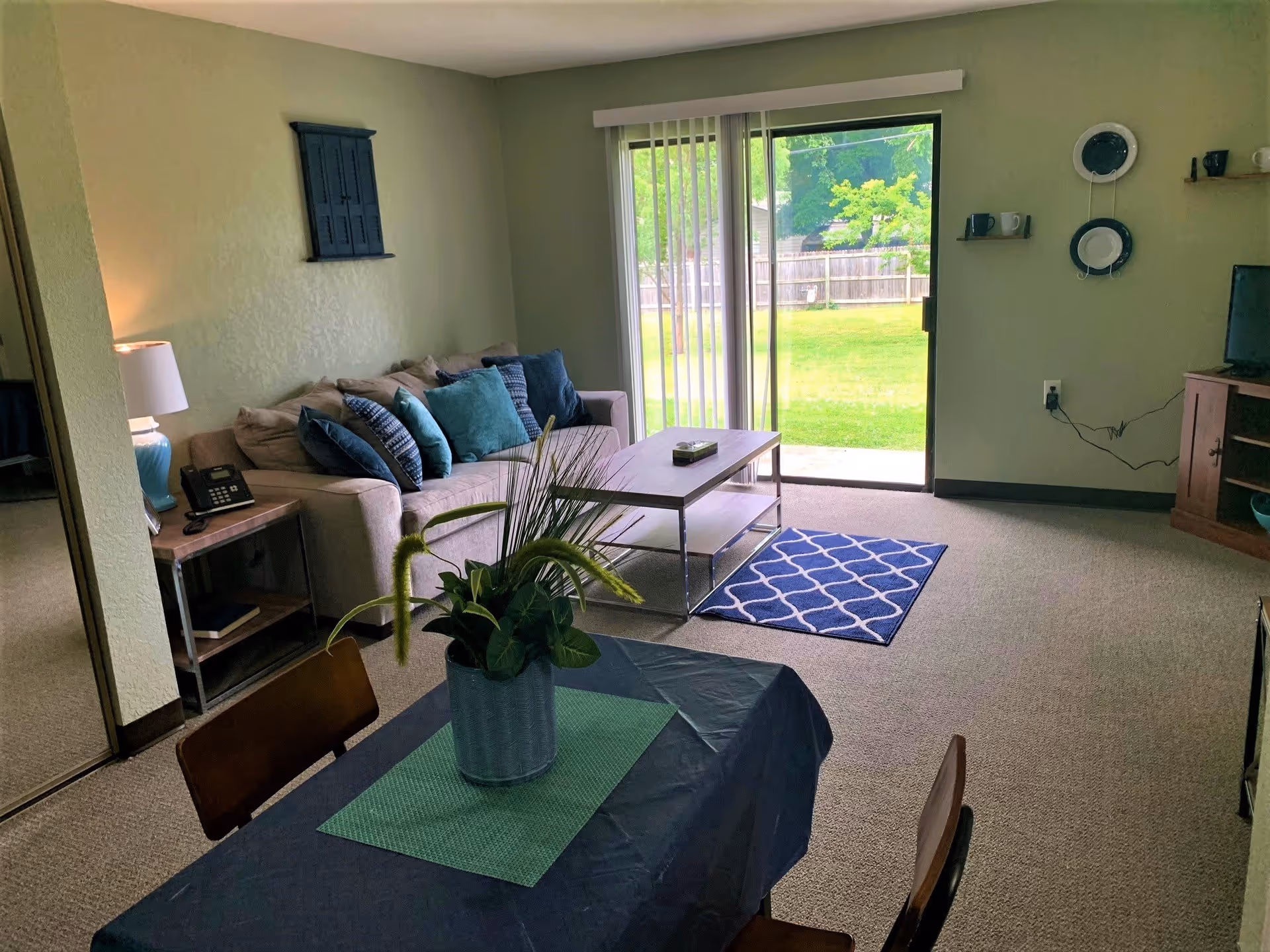 A cozy living room with a beige couch adorned with multiple blue and teal pillows, a wooden coffee table on a small blue patterned rug, and a sliding glass door leading to a green backyard. A small dining table with a blue tablecloth and a potted plant is in the foreground. The walls are light green with decorative plates and shelves, and a TV is placed on a wooden stand to the right.