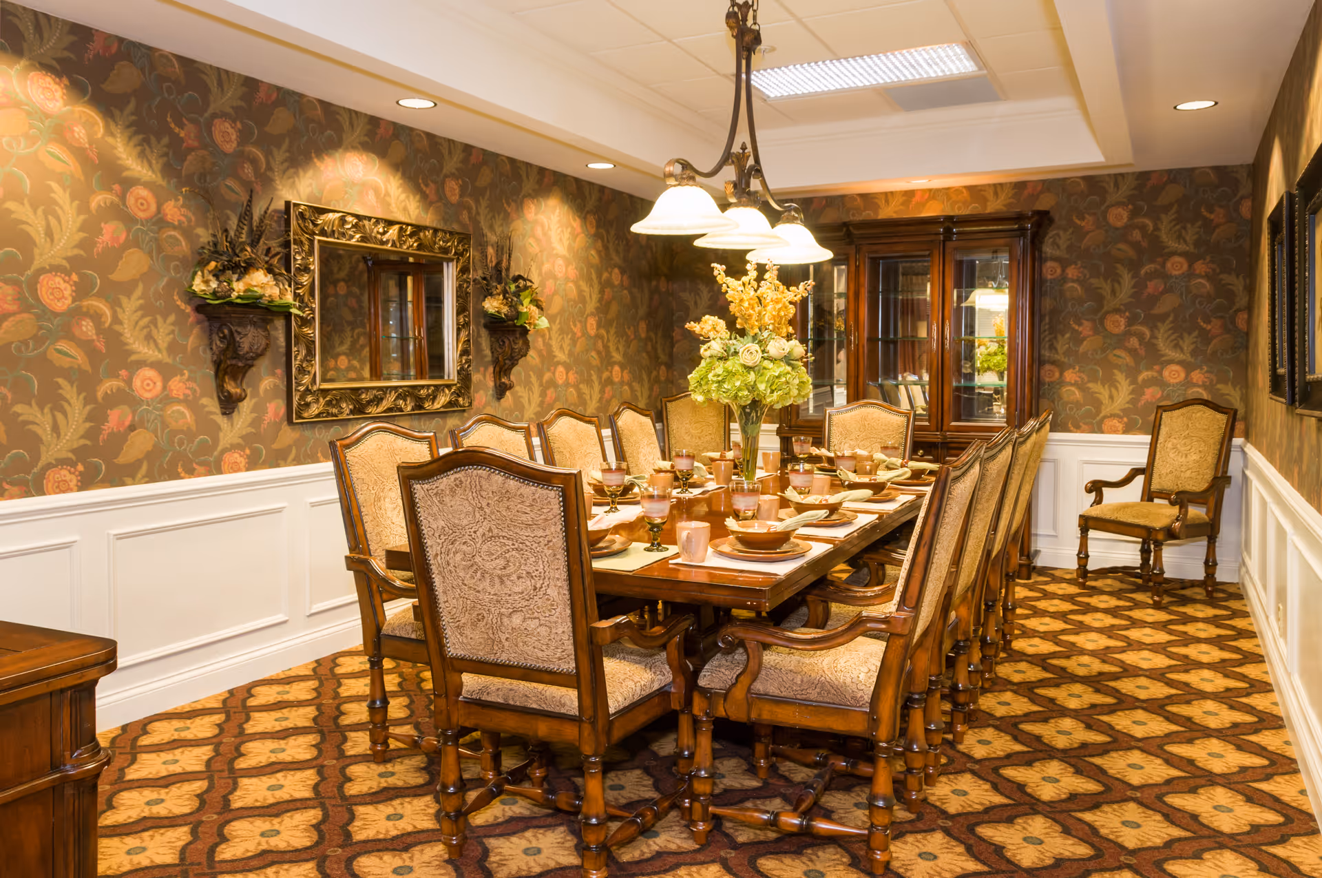 A formal dining room with a long wooden table set for a meal, surrounded by upholstered chairs. The room features floral patterned wallpaper, a decorative mirror on the wall, two wall-mounted flower arrangements, a chandelier with multiple lights hanging above the table, and a wooden china cabinet in the corner. The carpet has a geometric floral pattern.