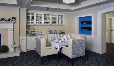 A cozy dining area with a square table set for a meal, surrounded by four upholstered chairs with patterned backs. The room features built-in cabinetry with glass doors displaying dishes, a fireplace with decorative items on the mantel, and a blue accent wall with two wooden shelves. The carpet has a geometric pattern, and the walls are painted in neutral tones.