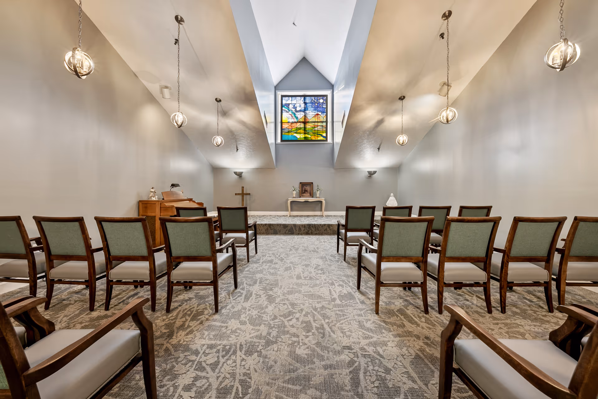 Interior view of a small chapel or meditation room with rows of wooden chairs with green cushions facing a raised platform. On the platform, there is a small table with religious items, a cross, and a stained glass window depicting a colorful landscape with mountains and a sun above.