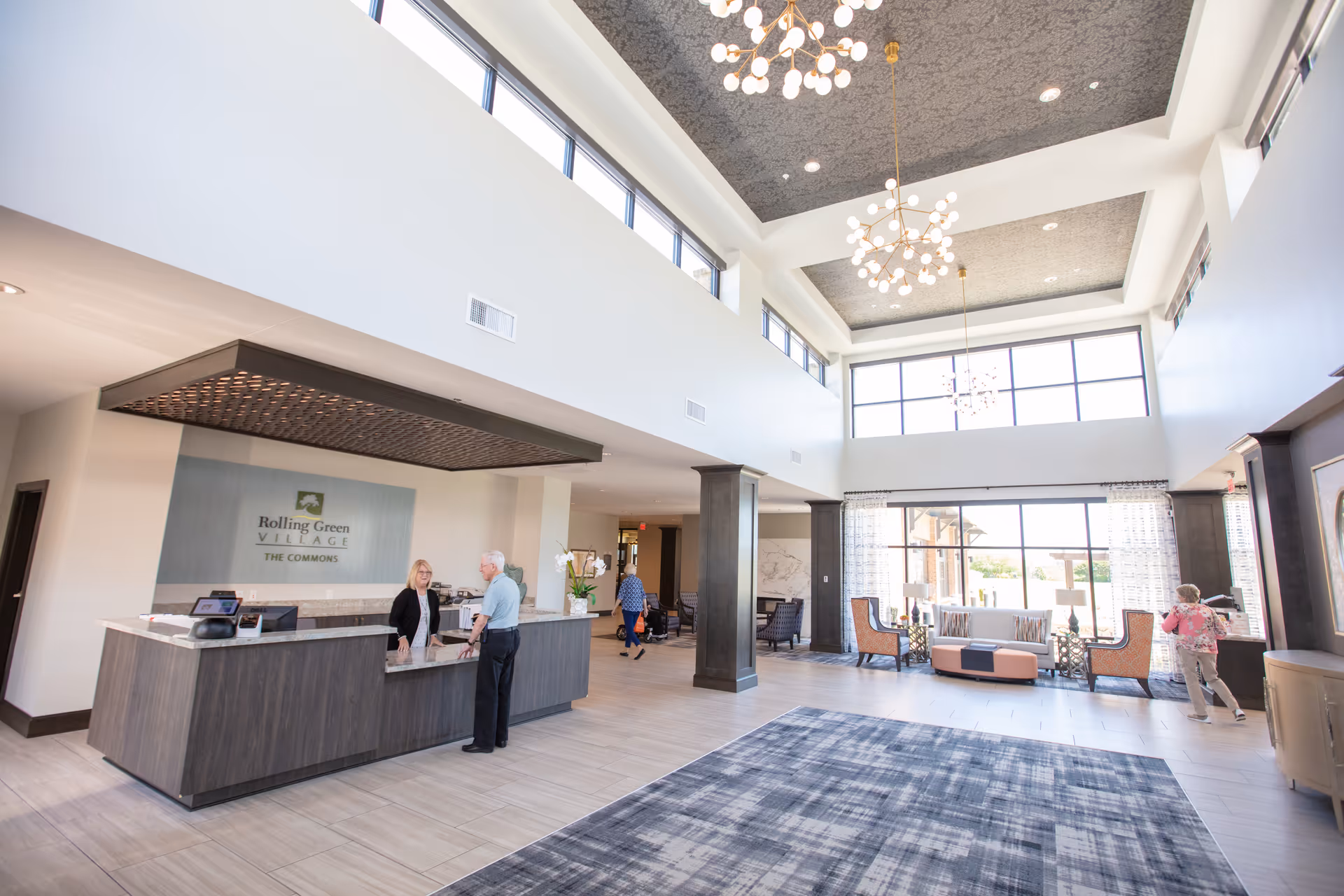 Spacious and bright senior living facility lobby with high ceilings, large windows, modern chandeliers, a reception desk with a staff member assisting an elderly man, and a seating area with chairs and a sofa near the windows.