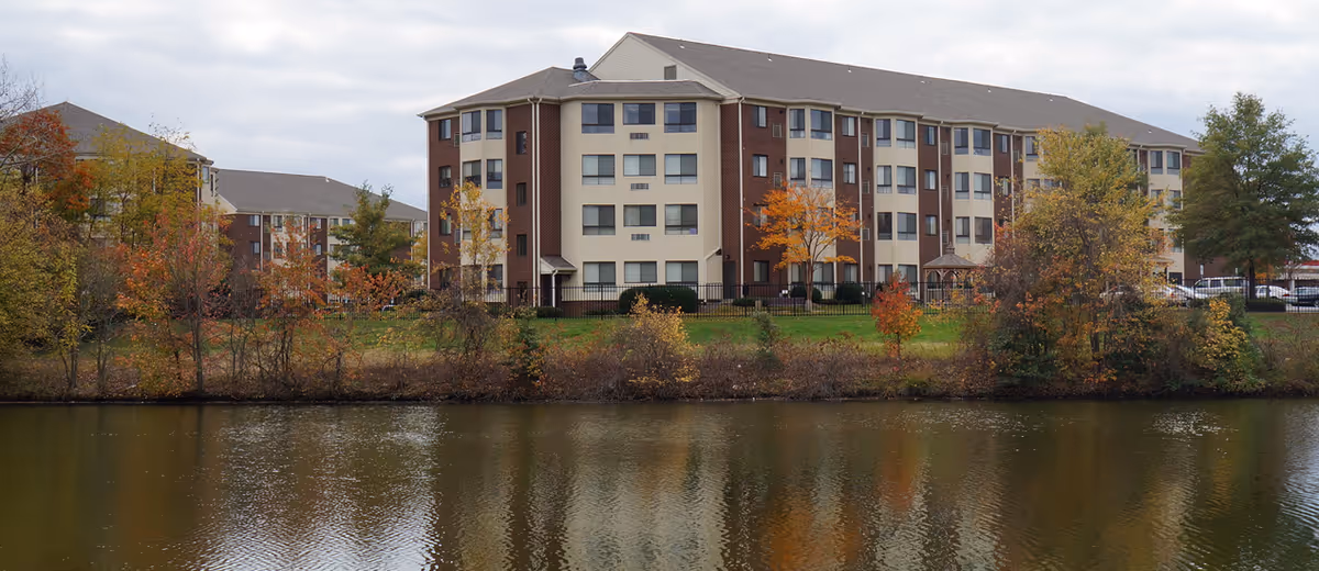 Multi-story brick and beige senior living building beside a lake with autumn trees and reflections in the water.