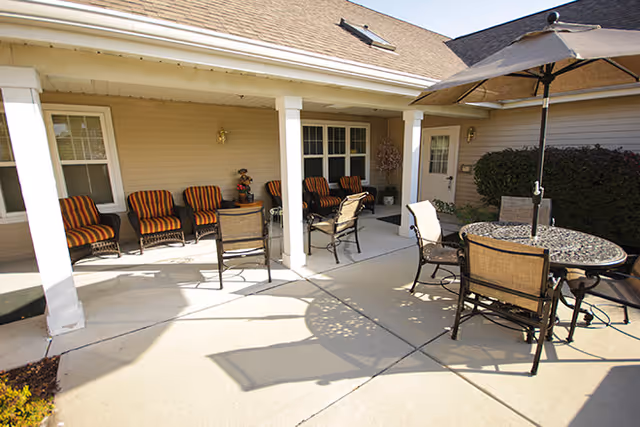 Covered outdoor patio with cushioned chairs, a round table with umbrella, and columns along the building.