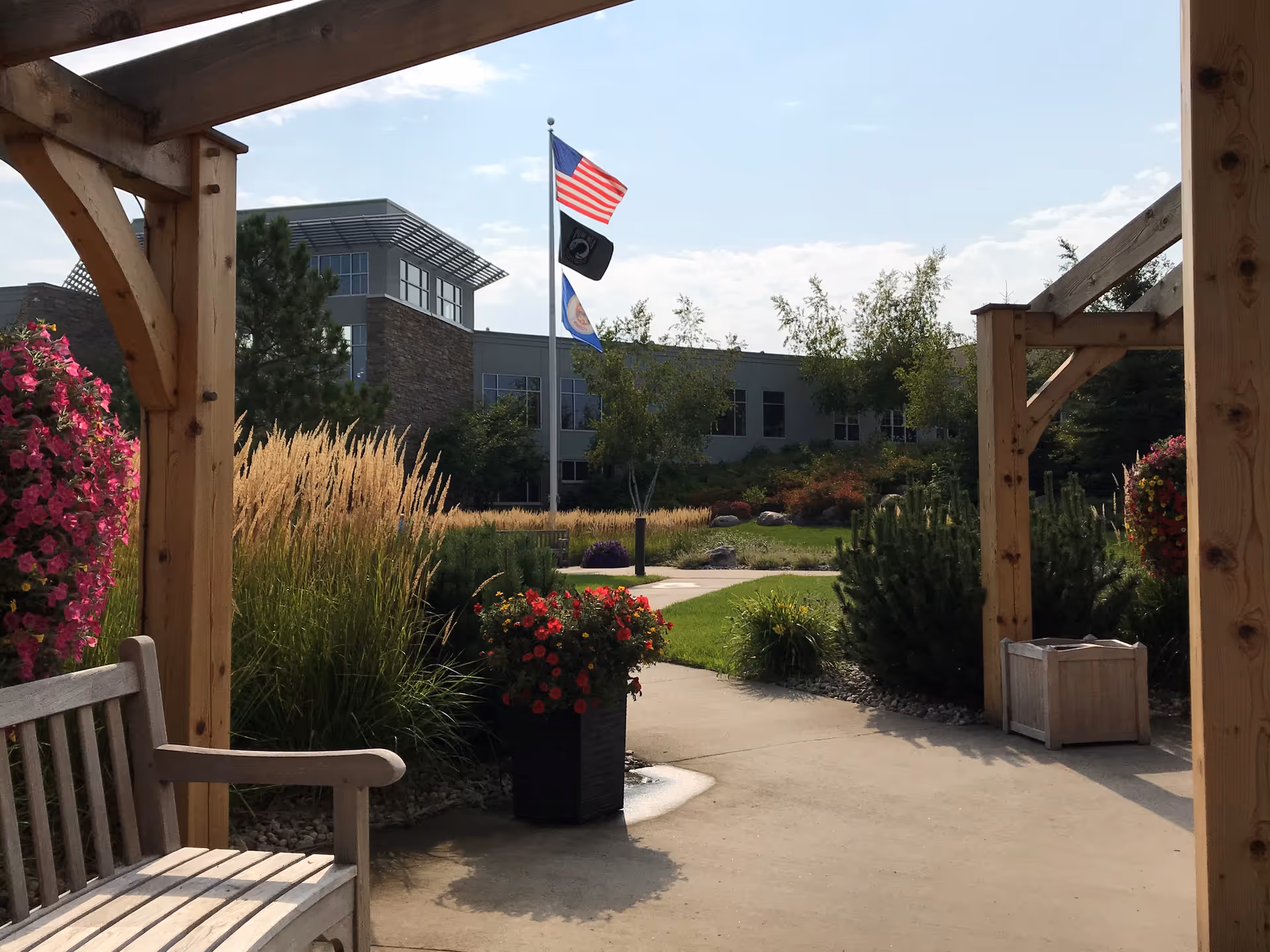View of an outdoor garden area at PioneerCare Center with a wooden pergola structure, a bench, flower pots with colorful flowers, ornamental grasses, and shrubs. In the background, there is a building with large windows and three flags flying on a flagpole under a partly cloudy sky.