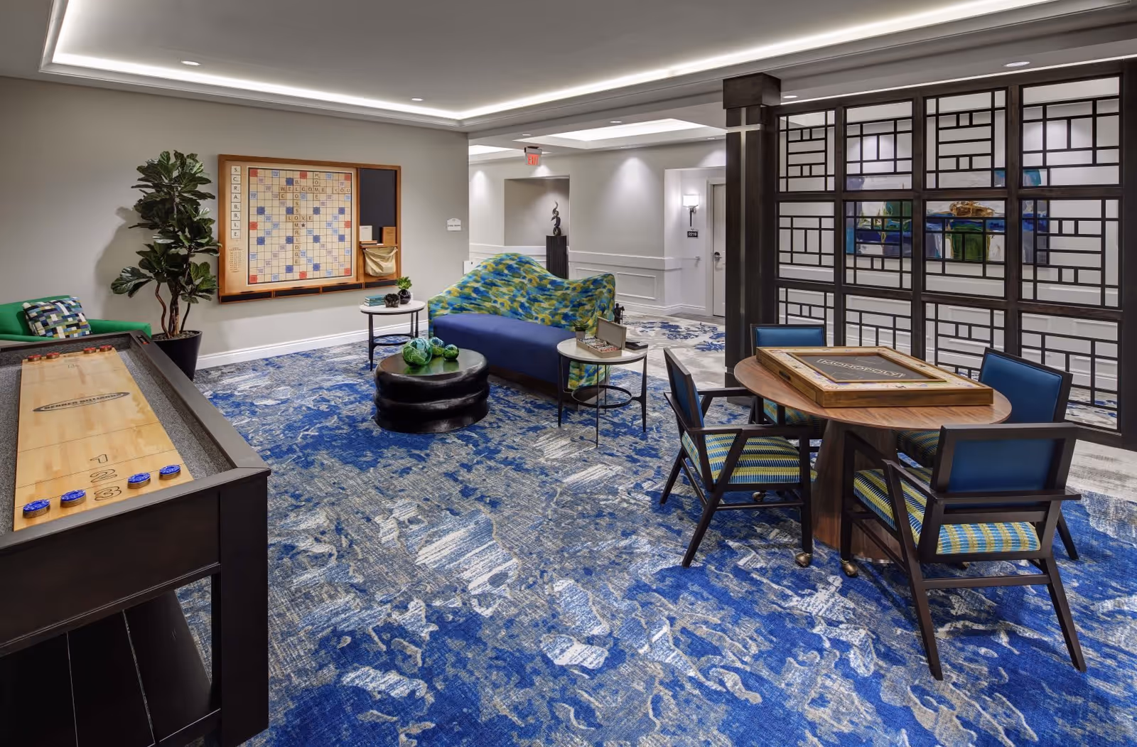 A recreational room with a blue patterned carpet featuring a shuffleboard table on the left, a large Scrabble board mounted on the wall, a blue and green patterned couch with a black ottoman and small round tables, and a round wooden table with four chairs. The room is well-lit with recessed lighting and has a decorative partition with geometric patterns on the right side.