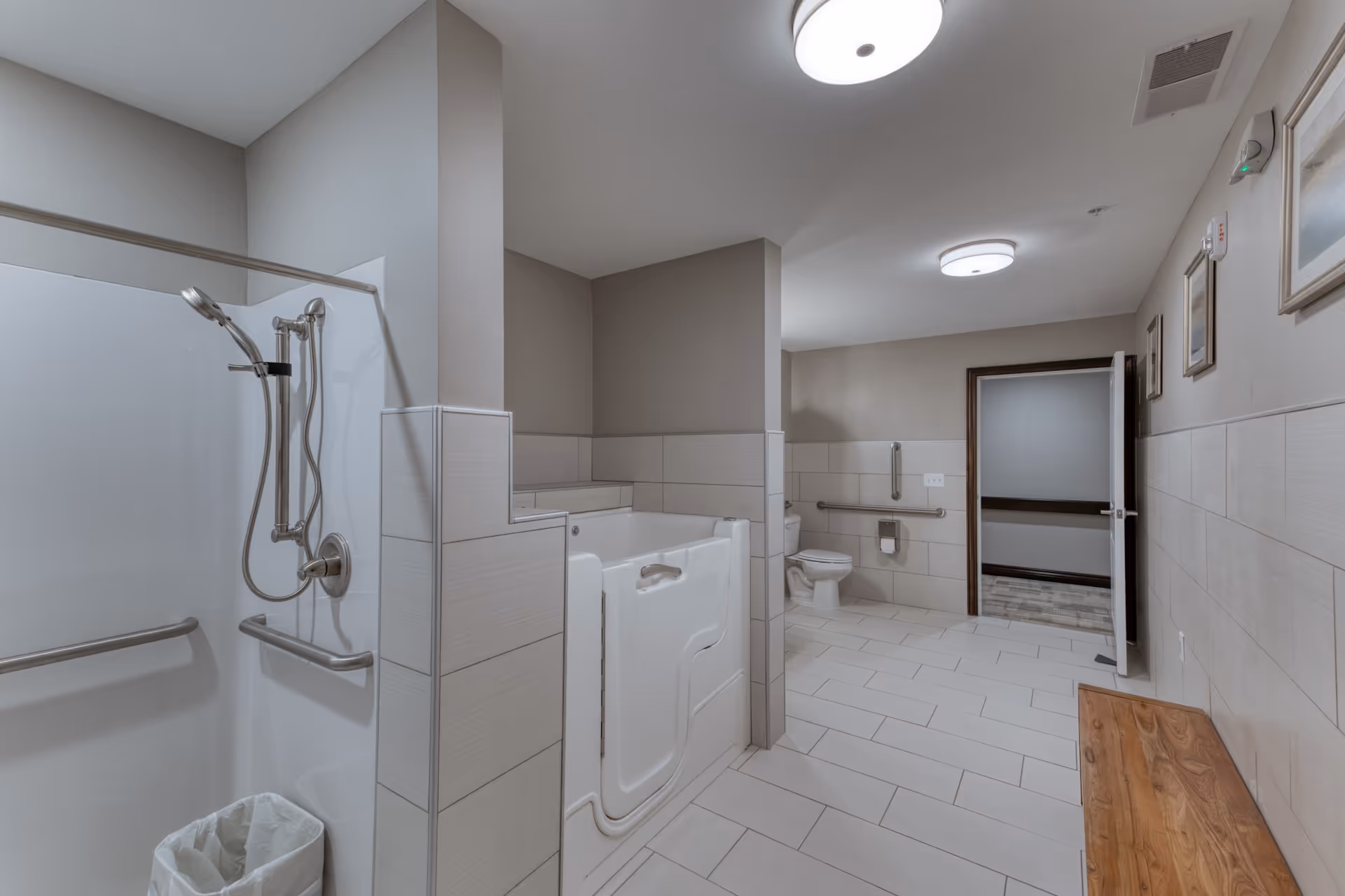 A spacious senior living bathroom featuring a walk-in bathtub with a door, a shower area with grab bars and a handheld showerhead, a toilet with a grab bar, light-colored tiled walls and floor, a wooden bench, framed pictures on the wall, and ceiling lights. The door to the bathroom is open, showing a hallway outside.