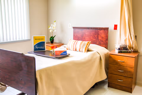 A neatly made single bed with a wooden headboard and footboard in a well-lit room. The bed has a beige bedspread and a decorative pillow. On the bed is a tray holding a welcome book and a small flower arrangement. Next to the bed is a wooden nightstand with a potted orchid and another wooden dresser with a stack of books. A window with vertical blinds and a curtain is visible, allowing natural light into the room.