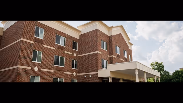 Three-story brick senior living building with a covered entrance and rows of windows under a partly cloudy sky.