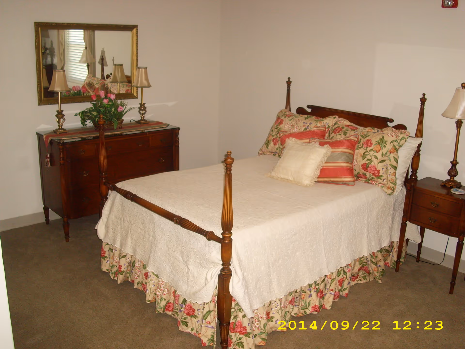 A bedroom with a wooden four-poster bed covered with a white quilt and floral bed skirt. The bed has multiple pillows with floral and striped patterns. To the left of the bed is a wooden dresser with two lamps and a vase of pink flowers, and a large mirror hanging above it. To the right of the bed is a wooden nightstand with a lamp. The room has beige carpet and plain white walls.