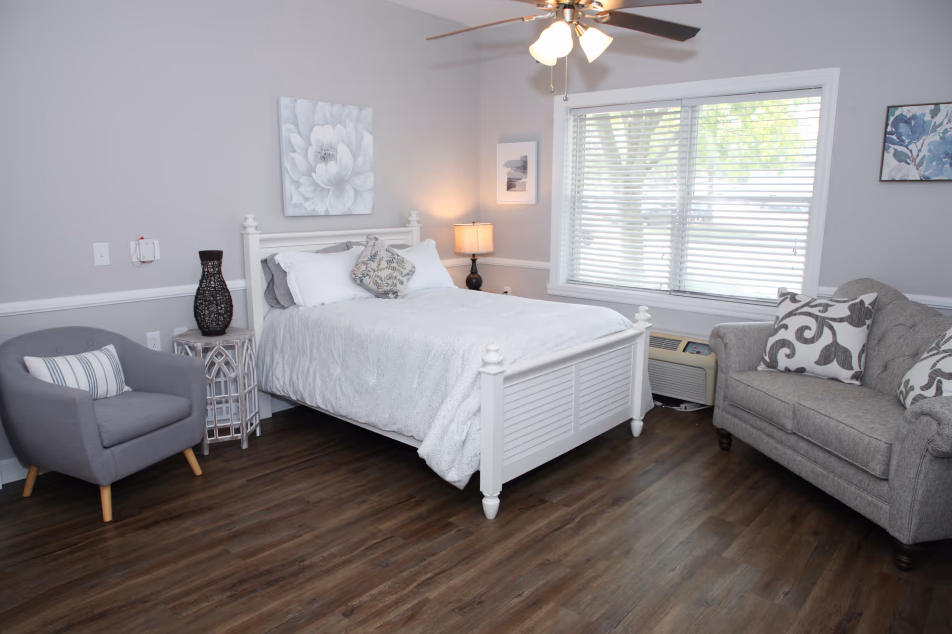 A cozy bedroom with a white wooden bed frame, white bedding, and decorative pillows. There is a gray armchair with a striped pillow to the left, a small side table with a dark vase, and a lamp on a nightstand next to the bed. A large window with blinds lets in natural light, and a gray couch with patterned pillows is positioned to the right. The room has wood flooring and light gray walls with floral artwork.