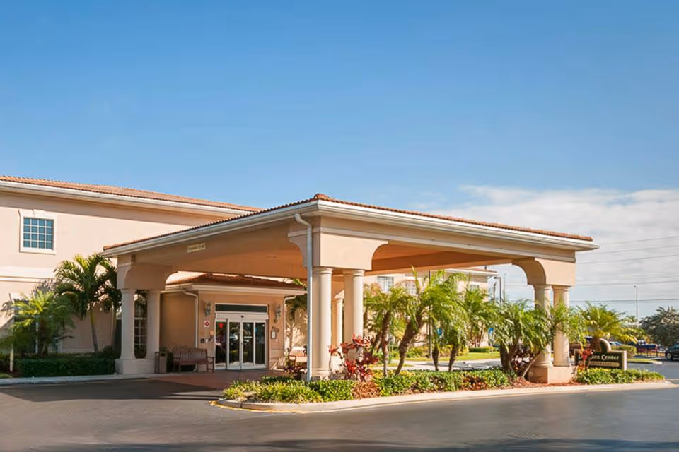 Entrance and covered porte-cochere of a senior living facility with palm trees and a driveway.