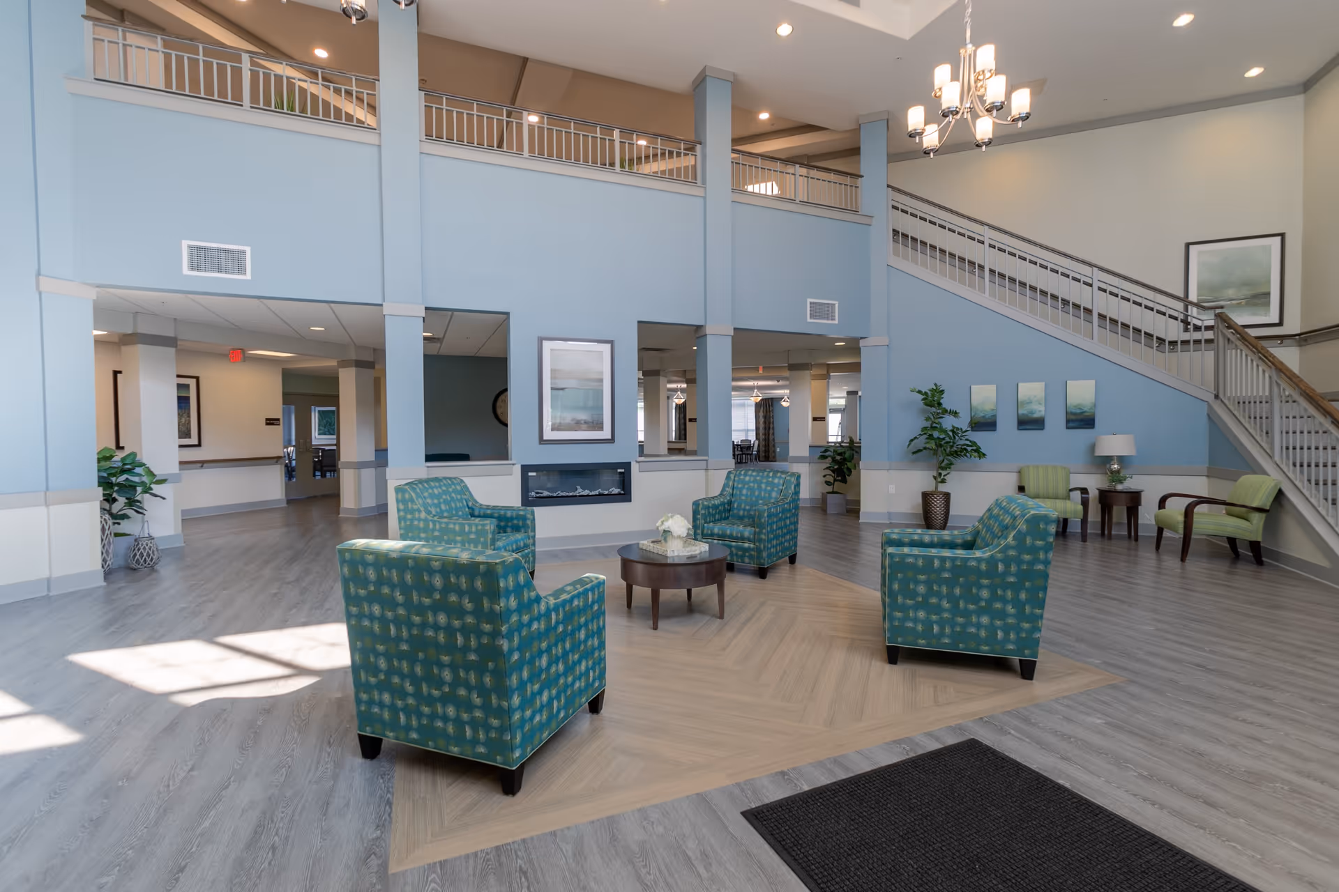 Spacious senior living facility common area with four teal patterned armchairs arranged around a round wooden coffee table on a light wood floor. The room features high ceilings, a modern fireplace, light blue walls, a staircase with railing, several framed artworks, potted plants, and a chandelier.