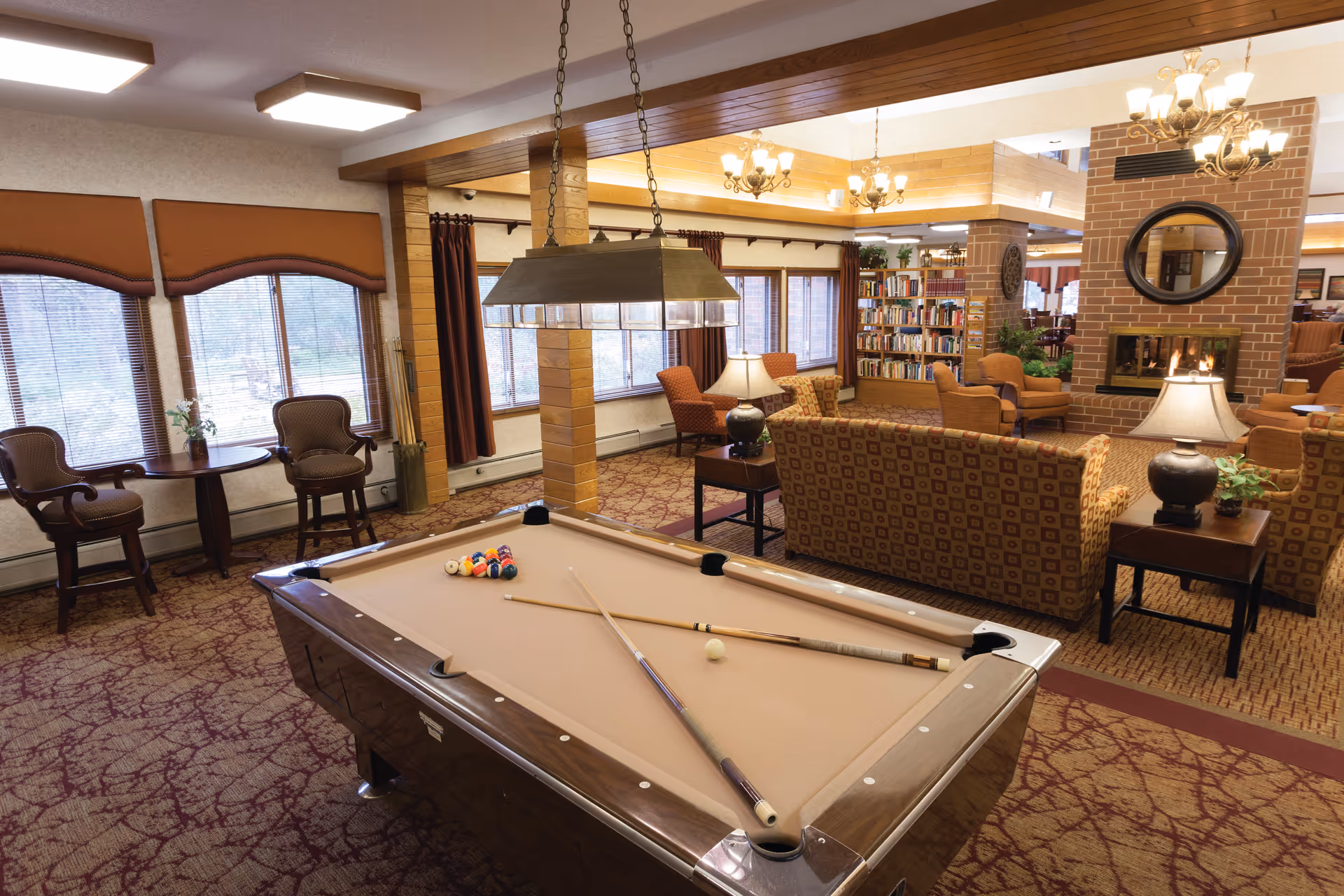 A cozy senior living common room with a pool table in the foreground and sofas, chairs, a fireplace and bookshelves in the background.