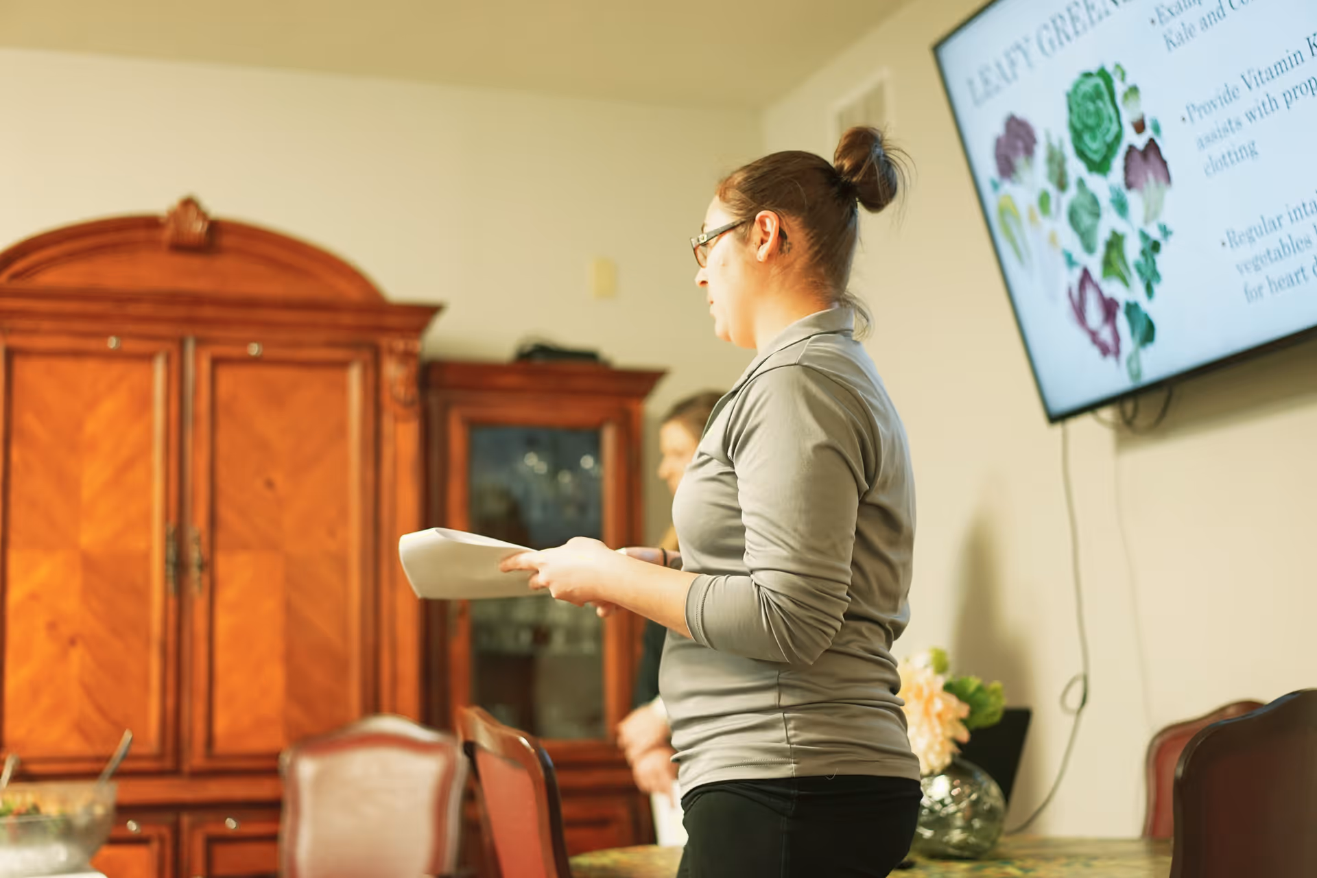 A woman standing in a room holding papers, with wooden cabinets and chairs around a table. A large screen on the wall displays information about leafy greens.