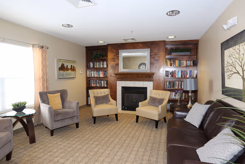 A cozy senior living room with a fireplace centered on a wooden wall unit filled with books. The room has two beige armchairs with gray pillows, a gray armchair with a yellow pillow, and a dark brown leather sofa with two light gray pillows. There is a glass side table with a small plant near the window with patterned curtains. The walls are decorated with framed artwork, and the floor is carpeted.