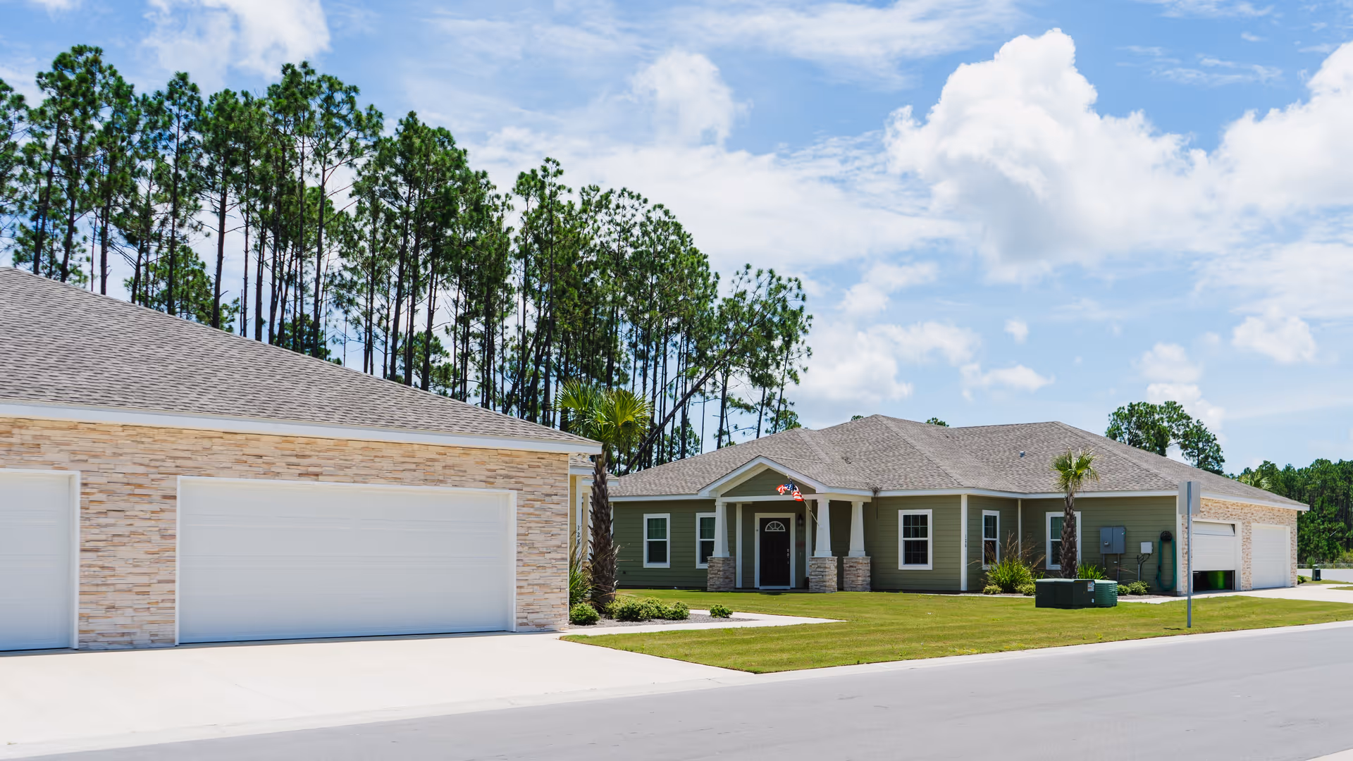 Single-story green residential building with attached garages, manicured lawn, palm trees and pine trees under a blue sky.