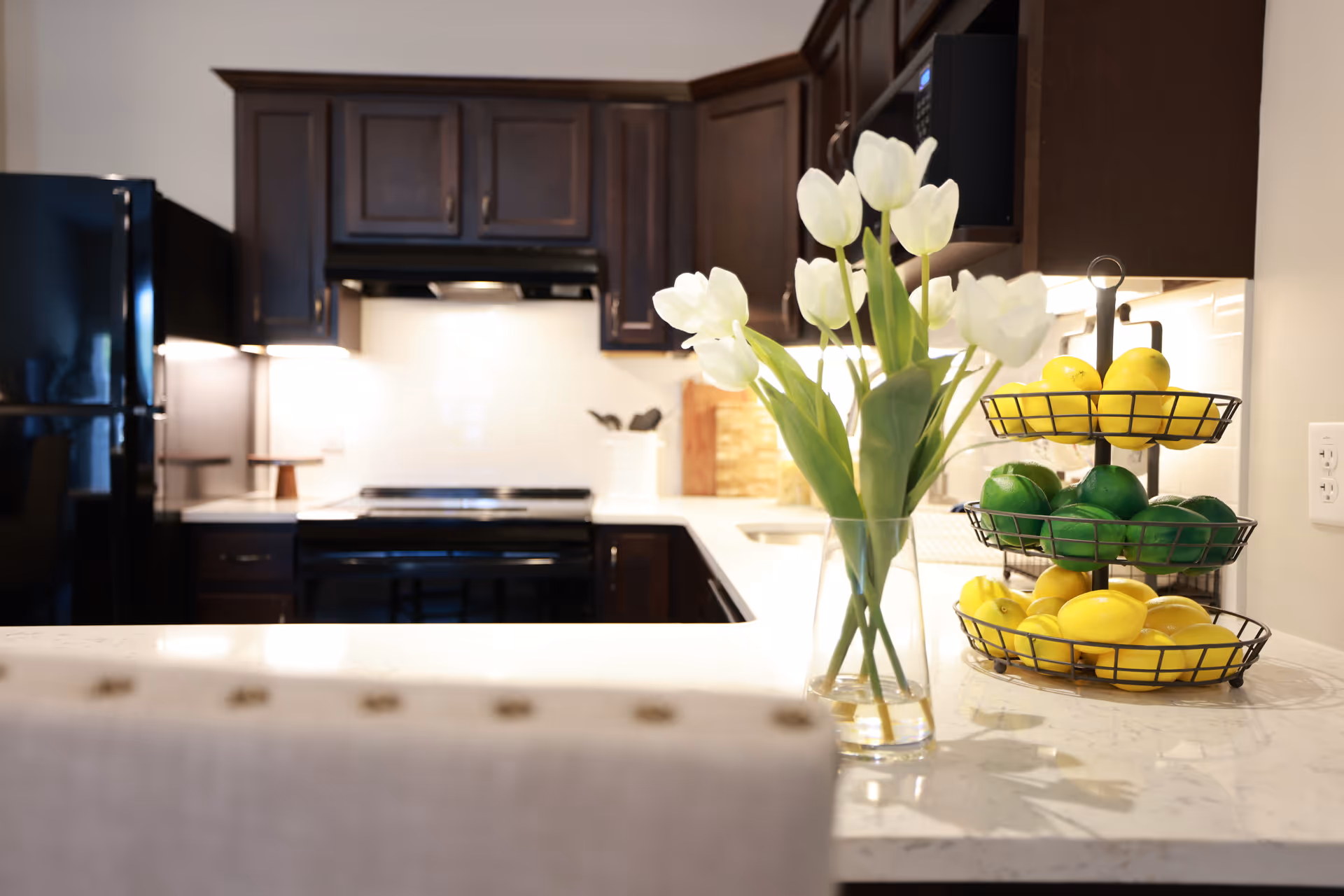 View of a modern kitchen countertop with a vase of white tulips and a three-tier fruit basket in front of dark cabinets and appliances.
