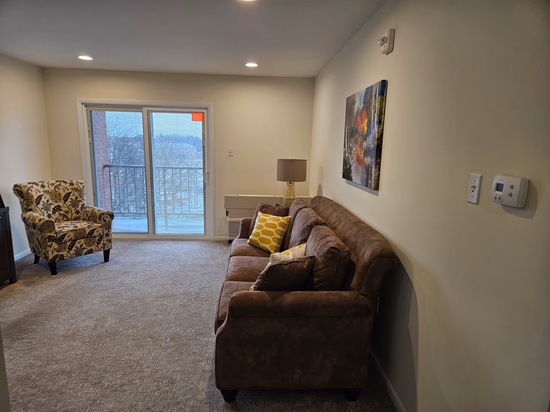 A cozy living room with a brown sofa adorned with yellow and brown pillows, a patterned armchair, a lamp on a side table, and a colorful painting on the wall. There is a sliding glass door leading to a balcony with a view of trees outside.