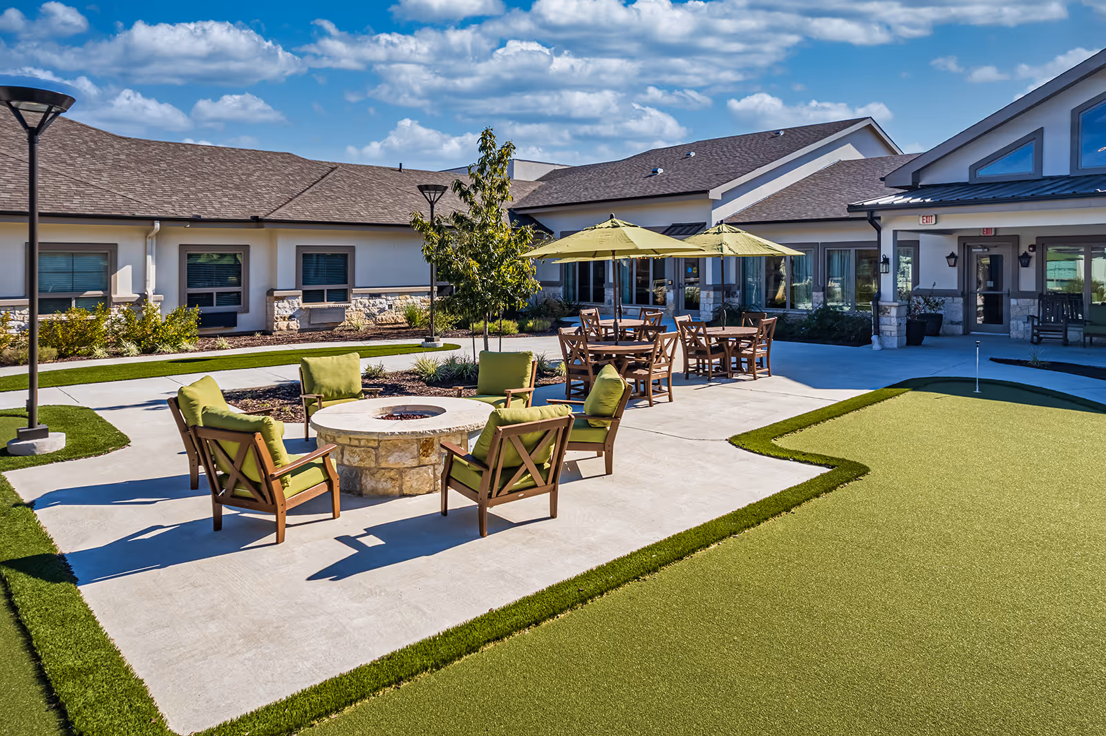 Outdoor patio area at Sage Valley Senior Living with green cushioned chairs arranged around a stone fire pit, wooden tables with green umbrellas, a small putting green, and a building with multiple windows and doors in the background under a partly cloudy sky.