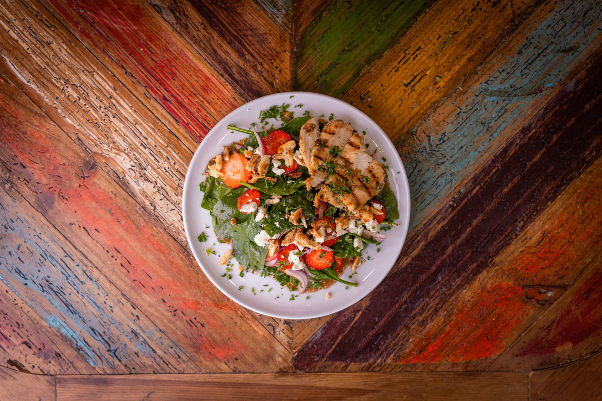 A white plate with grilled chicken and a spinach-strawberry salad on a colorful wooden table.