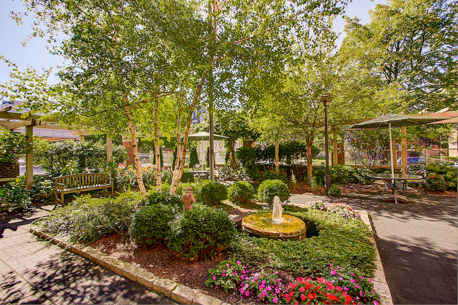 A lush outdoor garden area with green trees, bushes, and colorful flowers surrounding a small circular water fountain. There are benches and tables with umbrellas providing shaded seating areas along paved walkways.