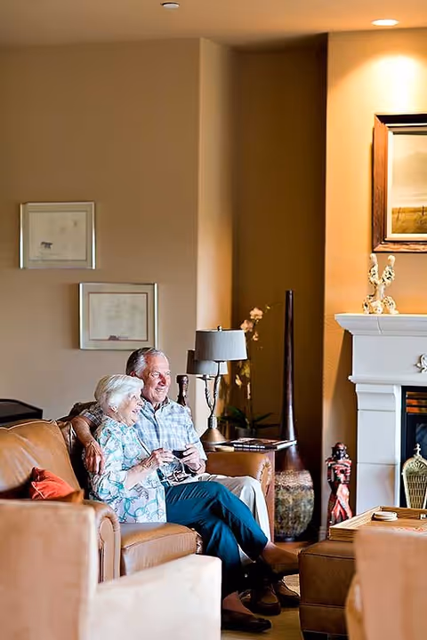 An elderly couple sitting closely together on a brown leather couch in a warmly lit living room. The man has his arm around the woman, and they both appear happy and relaxed. The room features a fireplace with decorative items on the mantel, framed pictures on the wall, a table lamp, and various decorative vases and sculptures.