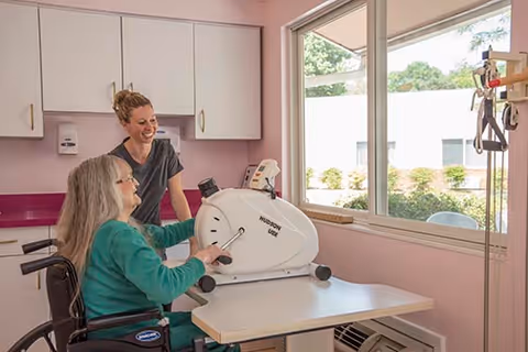 A woman in a wheelchair using a hand pedal exercise machine on a table near a window, assisted by a smiling caregiver standing beside her in a room with white cabinets and pink walls.