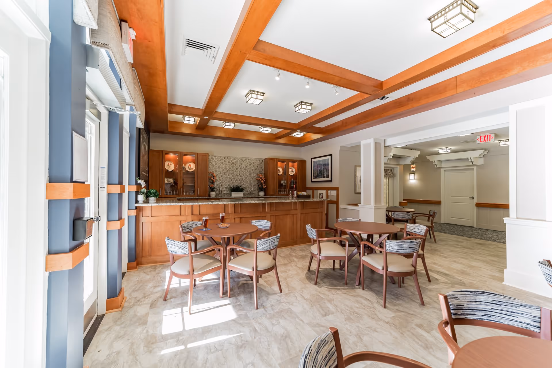 A bright and spacious common dining area in a senior living facility with round wooden tables and cushioned chairs. The room features a wooden coffered ceiling with modern light fixtures, a counter with cabinets and decorative items, and large windows letting in natural light. The flooring is light-colored tile, and there is an exit sign visible in the background.