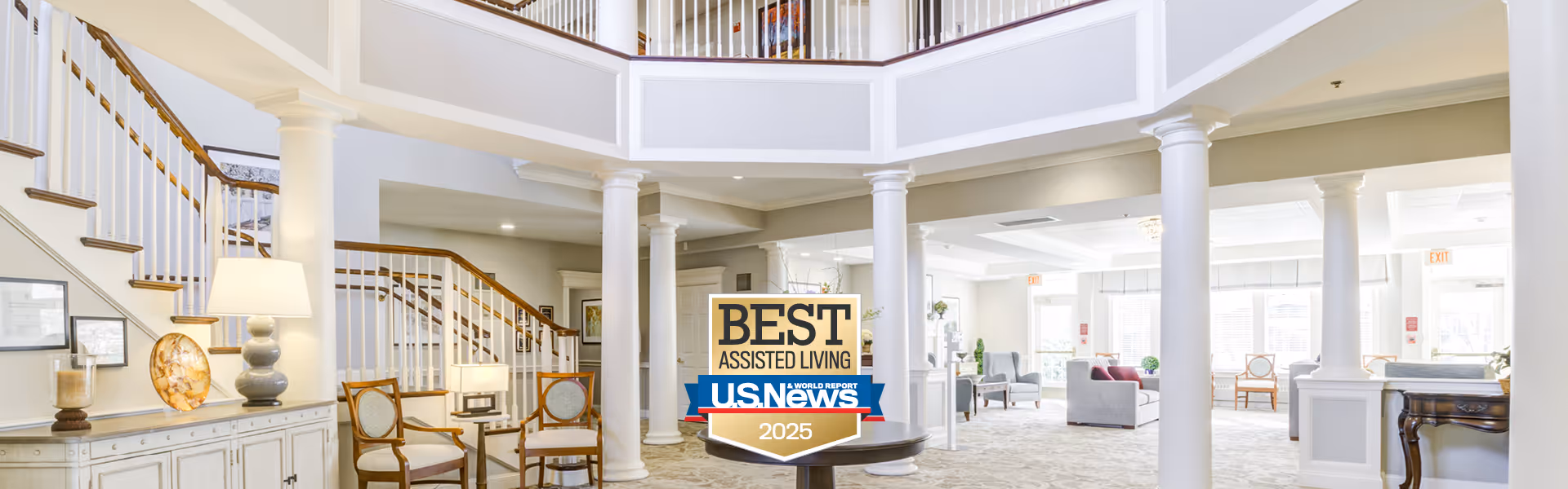 Bright and spacious senior living facility lobby with white columns, a staircase with wooden handrails, elegant chairs, a table with a lamp, and a large sign reading 'Best Assisted Living U.S. News 2025'.