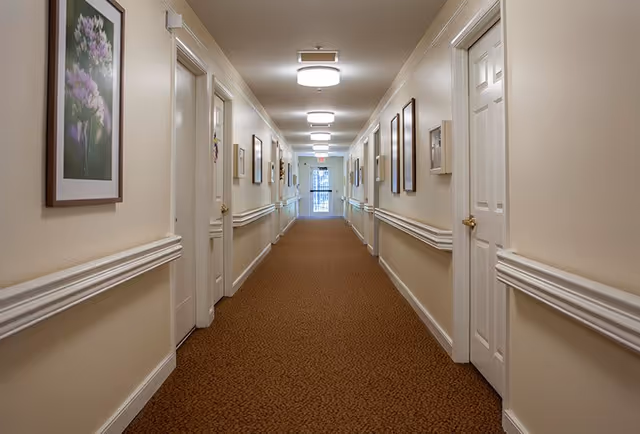 A long, well-lit hallway in a senior living facility with beige walls, brown carpet, white doors on both sides, handrails along the walls, and framed artwork hanging on the walls.