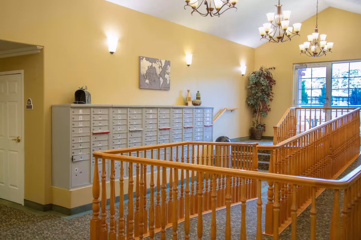 Interior hallway area with a set of mailboxes mounted on a yellow wall, a wooden railing surrounding a stairwell, a potted plant in the corner, and large windows letting in natural light. The space is lit by wall sconces and chandeliers hanging from the ceiling.