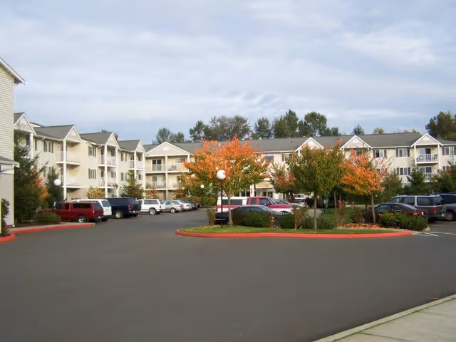 Parking lot area in front of a multi-story senior living facility building with several cars parked and trees with autumn foliage in landscaped islands.