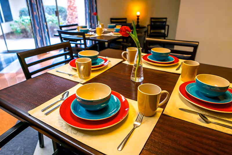 A dining table set for four with colorful plates, bowls, and mugs. Each place setting has a red plate, a blue bowl, a beige mug, and silverware on cream-colored placemats. A small glass vase with a single red flower is in the center of the table. The background shows more tables and chairs near a window with curtains.
