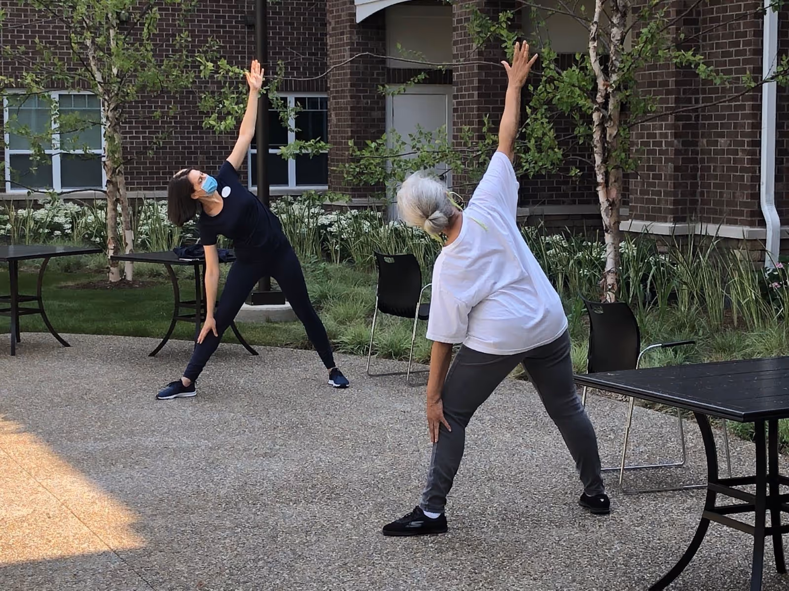 Two women, one younger and one older, are outdoors on a patio area in front of a brick building. They are both stretching with one arm reaching up and the other touching their leg. The younger woman is wearing a face mask, black outfit, and sneakers, while the older woman is wearing a white t-shirt, gray pants, and black shoes. There are black tables and chairs around them, and greenery including small trees and plants near the building.