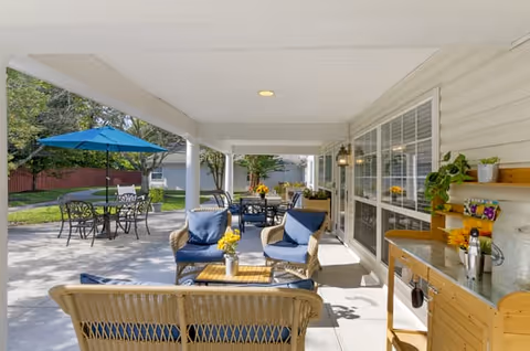 Covered outdoor patio area with wicker seating including chairs and a loveseat with blue cushions, a small table with a vase of yellow flowers, and additional tables and chairs with a blue umbrella in the background. The patio is adjacent to a building with large windows and light-colored siding, surrounded by green grass and trees.