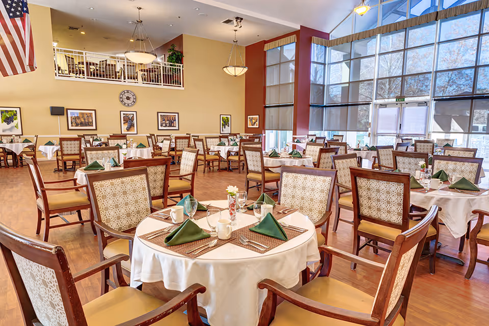 A spacious dining room with multiple round tables covered with white tablecloths, each set with green folded napkins, cups, glasses, and silverware. The room has large windows with roller shades, wooden flooring, and beige walls adorned with framed pictures and a clock. There is an American flag hanging on the left side and a mezzanine level with additional seating.