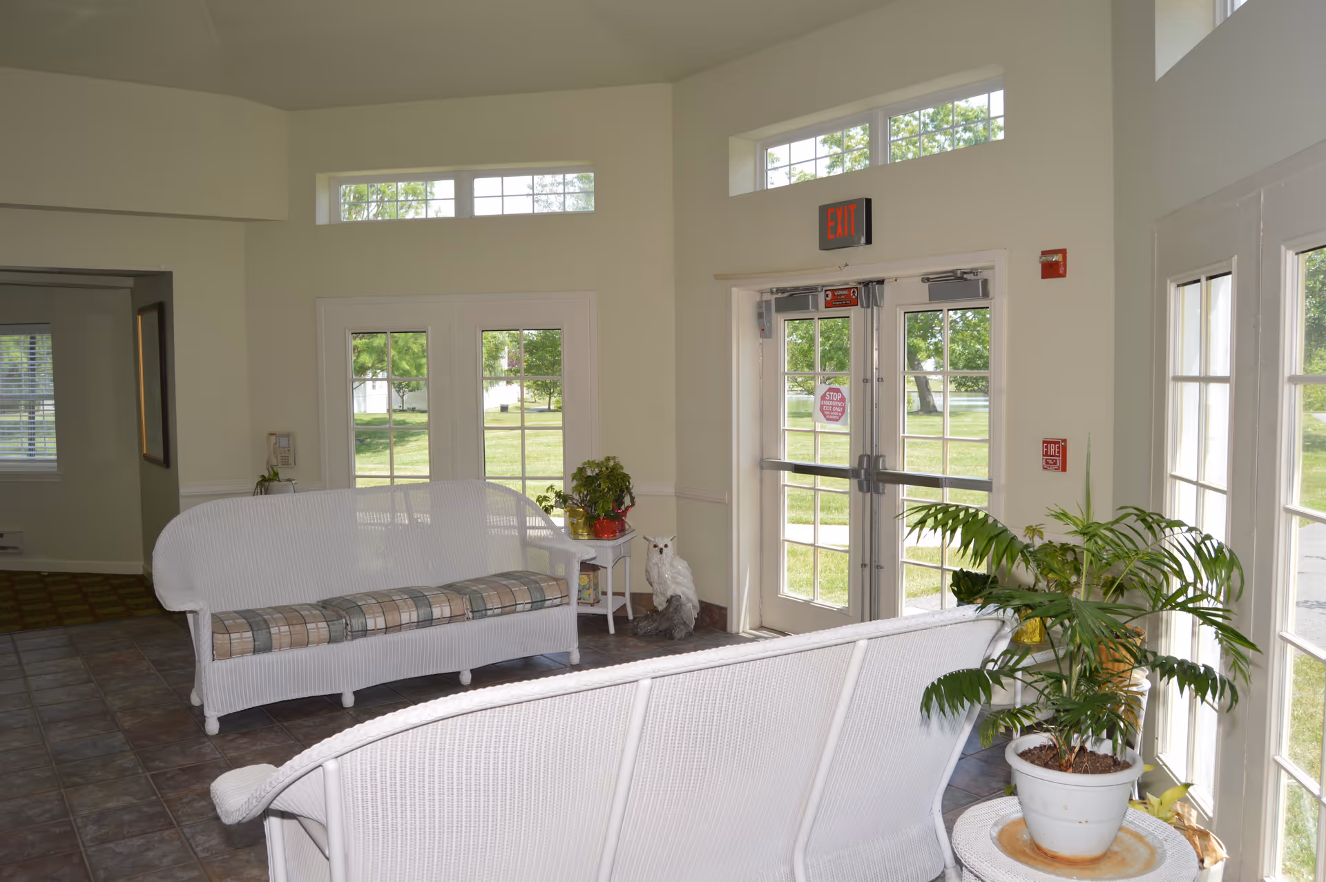 A bright and airy interior seating area with white wicker furniture including two sofas with plaid cushions. There are large windows and glass doors letting in natural light, potted plants on a small table and on the floor, and an exit sign above the door. Outside, green grass and trees are visible through the windows.