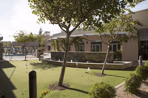 Outdoor view of a senior living facility courtyard at Beatitudes Campus, featuring a putting green with small flags, trees, shrubs, and a building with large windows in the background under a clear sky.