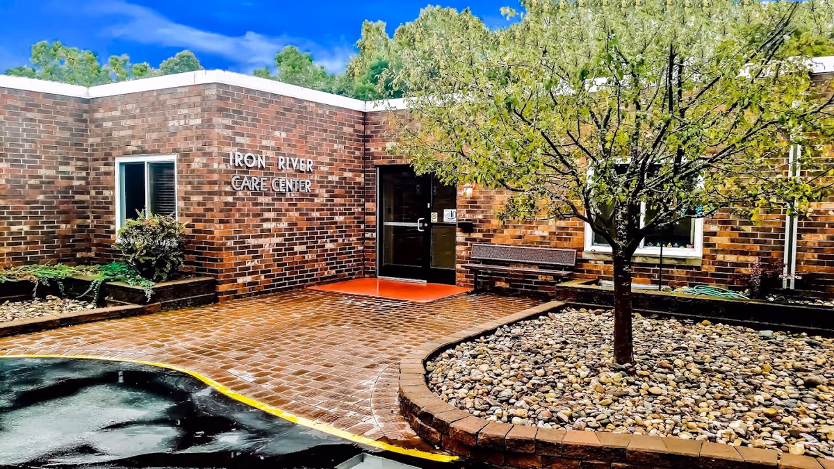Exterior view of the Iron River Care Center building entrance with a brick facade, a glass door, a bench, a tree planted in a circular stone bed, and a paved walkway.