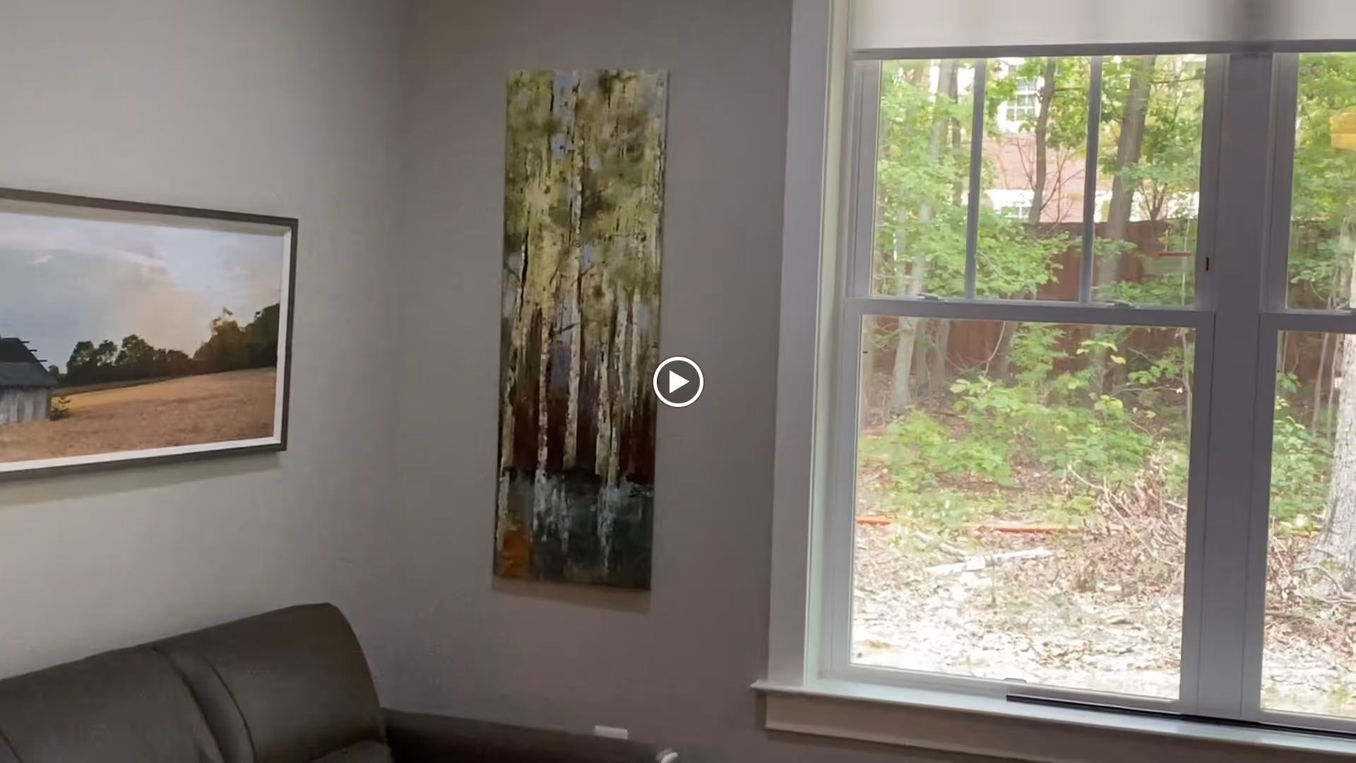 Corner of a living room with a sofa, two pieces of wall art, and a large window looking out to trees.