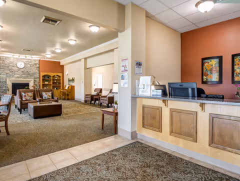 Interior view of a senior living facility lobby with a reception desk on the right, decorated with two framed floral paintings above it. The lobby area has carpeted flooring, several chairs and ottomans arranged around a fireplace with a stone surround and a clock above it. There are bookshelves and additional seating in the background, with warm lighting from ceiling fixtures.