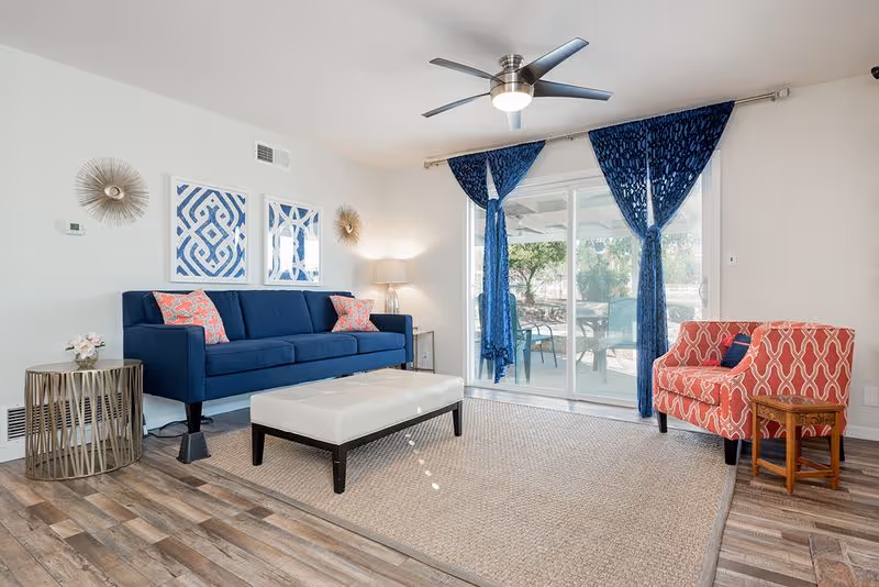 A bright living room with a blue sofa adorned with pink patterned pillows, a white cushioned ottoman, and a red patterned armchair next to a small wooden side table. The room features wood flooring, a beige area rug, blue curtains tied back on a sliding glass door leading to an outdoor patio with chairs and a table. Wall decorations include two framed blue and white geometric prints and two sunburst wall sculptures. A floor lamp is placed in the corner near the sofa.